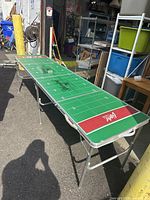Full view of folding beer pong table with green football field design and Yuengling branding. Metal frame and foldable legs visible.
