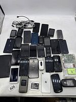 Photo showing a collection of various cell phones, a tablet, cables and circuit boards on a white surface.