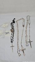 Five rosaries displayed on a white surface, showing beads and crosses with a bidding tag in the background.