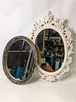 Both mirrors shown side by side against a plain background. White frame mirror is larger and has detailed ornamental design. Silver oval mirror is smaller with visible frame damage.