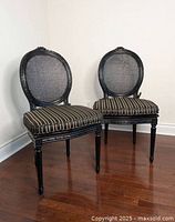 Photo showing two black solid wood Louis style chairs with cane backs and black/gold striped cushions sitting on a hardwood floor against a wall.
