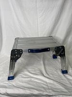 Front angled view showing aluminum platform, black metal joints, blue rubber feet, and black handle on one side.