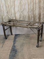 Side view of antique folding wooden washtub stand showing rectangular frame with slatted top and metal support braces, set on carpet with lace curtain backdrop.