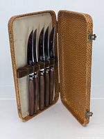 Set of five curved cutting utensils with wooden handles inside textured tan case with spotted pattern. Case is open showing utensils secured by wooden strip.