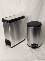Front and side view of two stainless steel trash cans on white backdrop: one rectangular and one smaller round shape with black lids and foot pedals.