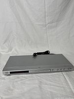 Front view of silver JVC DVD/CD player with power cord placed on top, showing front panel with buttons and disc tray.