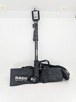 Photo showing the selfie stick extended vertically with a smartphone clamp and foam grip, along with the carrying case laid flat with Rado Switzerland branding.
