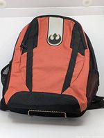 Front view of orange and black Star Wars backpack displaying Rebel Alliance insignia on top front pocket; two vertical zipper pulls on front pocket; mesh side pockets visible; black bottom and trim