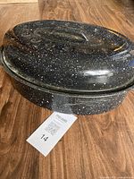 Photo of the enamelware roasting pan with lid on a wooden floor. The pan is black with white speckles and an attached handle on the lid.