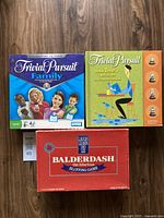 Three board games arranged side-by-side on a wooden floor: Trivial Pursuit Family, Trivial Pursuit Book Lover's Edition, and Balderdash 2nd Edition.