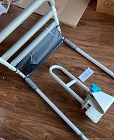 View of white metal bed rail frame with gray fabric storage pouch and separate white metal bath handle featuring blue adjustment clamp.