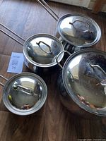 Top view of all four saucepans with their matching stainless steel lids on a hardwood floor, reflecting light and surroundings.