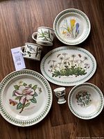 Full set of Portmeirion Botanical Garden ceramic dishware including plates, cups, egg cup, and bowl with floral butterfly designs arranged on wooden floor
