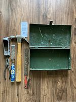 Green metal toolbox open showing interior and displayed tools including hammer, pliers, chisel with blue handle, and file