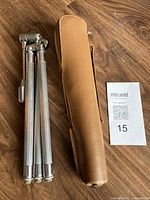 Folded Velbon metal camera tripod with chrome finish standing beside tan soft case with zipper closure laid on wood surface.