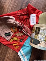 Photo showing three neck scarves (red, pink, blue), two badges, two pendants, a leather scarf closure, and a collectible tin