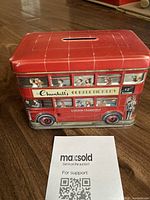 Front and side view of red tin money box shaped like a London double decker bus with vintage style illustrations of bus passengers and advertisements.