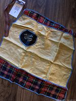 Full view of the vintage small apron laid flat on a wooden surface showing yellow rayon fabric with red-black tartan trim and an embroidered patch.