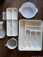 Five white kitchen items laid out on wooden floor including plastic trays, ceramic spoon, mug, and colander bowl.