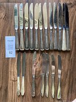 19 vintage silverplate table knives arranged in a row on wooden surface showing different handle patterns and blade shapes with visible tarnish and wear.