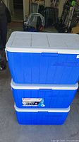 Stacked view of two blue Coleman coolers with white lids in a garage, showing exterior condition and brand