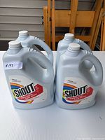 Four gray plastic bottles labeled Shout Triple Acting stain remover, each 3.8L, shown from front angle on white table outdoors.