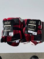 Two folded red and black buffalo check patterned Couture throw blankets with tags displaying product details and branding.