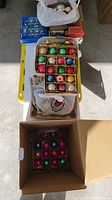 Top view showing multiple boxes containing vintage glass Christmas ornaments mostly round in shape, featuring diverse colors such as red, green, gold, blue, silver, pink, and yellow. Ornaments are packed in cardboard boxes with dividers and some loose ornaments visible.