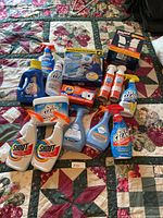 Photo showing full collection of cleaning supplies arranged on quilted bedspread, various bottles, sprays and boxed items.