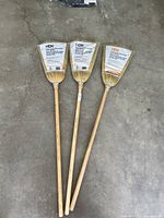 Three brand new HDX corn brooms laid out on concrete floor, showing full length wooden handles and natural broomcorn bristles with labels attached.