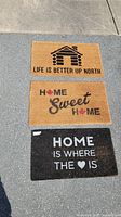 Three front entrance mats laid out on pavement showing text and designs.