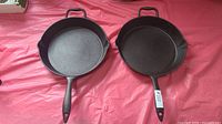 Two like-new 12-inch cast iron pans side by side on a red surface, showing their size and shape.