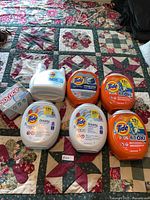 Six containers of Tide PODS and one large jug of Tide liquid detergent with one box of Bounce dryer sheets displayed on quilted bedspread.