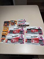 Photo showing various new batteries including Energizer Max AAA (16-pack), AA (10-pack), C and D size 2-packs, and Duracell Procell AA and AAA packs arranged on a table.