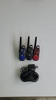 Three Uniden two-way radios in blue, black, and red positioned upright behind a black dual charging dock with power adapter on a white surface.