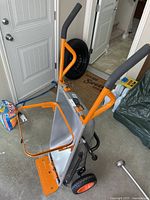 Front angle view showing wheelbarrow bucket, orange metal frame, black wheels, and handles with foam grips.