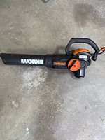 Full side view of Worx Trivac electric leaf blower on concrete, showing black and orange housing and Worx logo on blower tube.