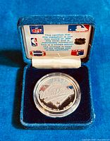 Silver medallion in clear protective capsule resting in a blue velvet display clamshell case, with certificate of authenticity showing logos of several sports leagues.
