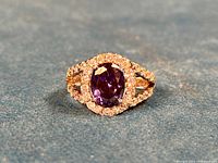 Close-up photo of ring lying flat on surface, showing oval purple amethyst CZ center stone surrounded by clear CZ stones and split shank design.