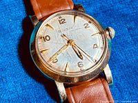 Close-up of watch face showing Bulova branding, gold-tone numerals and hands, light-colored dial with patina.