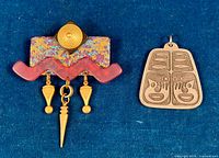 Top down view of both brooches on dark blue fabric background, showing colorful acrylic brooch with gold-tone dangles and engraved pewter brooch beside it.