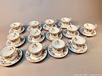 Complete set of 13 Royal Albert Brigadoon tea cups each paired with its matching saucer, displayed in rows on a flat surface. Both cups and saucers have visible blue and brown floral patterns with gilded rims.
