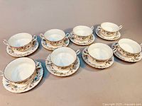 Set of 8 double handled soup bowls each paired with matching underplate arranged in 2 rows on a beige surface.