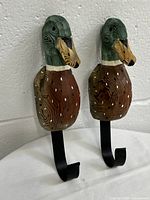 Front view of two vintage carved wooden duck head clothes hooks showing detailed painting and metal hooks beneath.