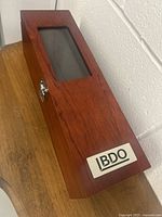 Photo of the wooden wine accessory box closed, showing the glass top panel, smooth reddish wood finish, metal clasp, and BDO metal branding plate.