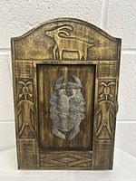 Front view of wooden key cabinet with carved goat and floral designs and metal figurative relief in center.