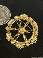 Front view of round lacework brooch with gold-tone metal and faux pearl accents next to a quarter for size reference.