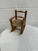 Front-facing photo of vintage wooden doll rocking chair with woven rope seat on white cloth background.