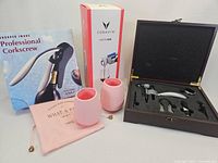 Photo showing Coravin Model One in open wooden box with accessories, Sharper Image professional corkscrew box, two pink silicone cups, and pink drawstring pouch.