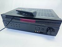 Front and top view of black Insignia AV digital home theater receiver with remote control resting on top, showing front buttons, headphones jack, input ports and volume knob.